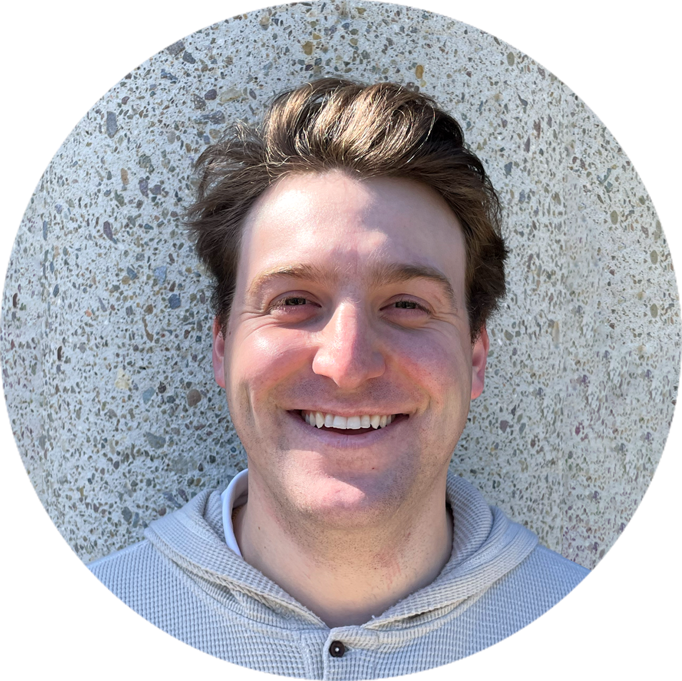 headshots-chris A person smiling, standing against a textured wall, wearing a light-colored hoodie.