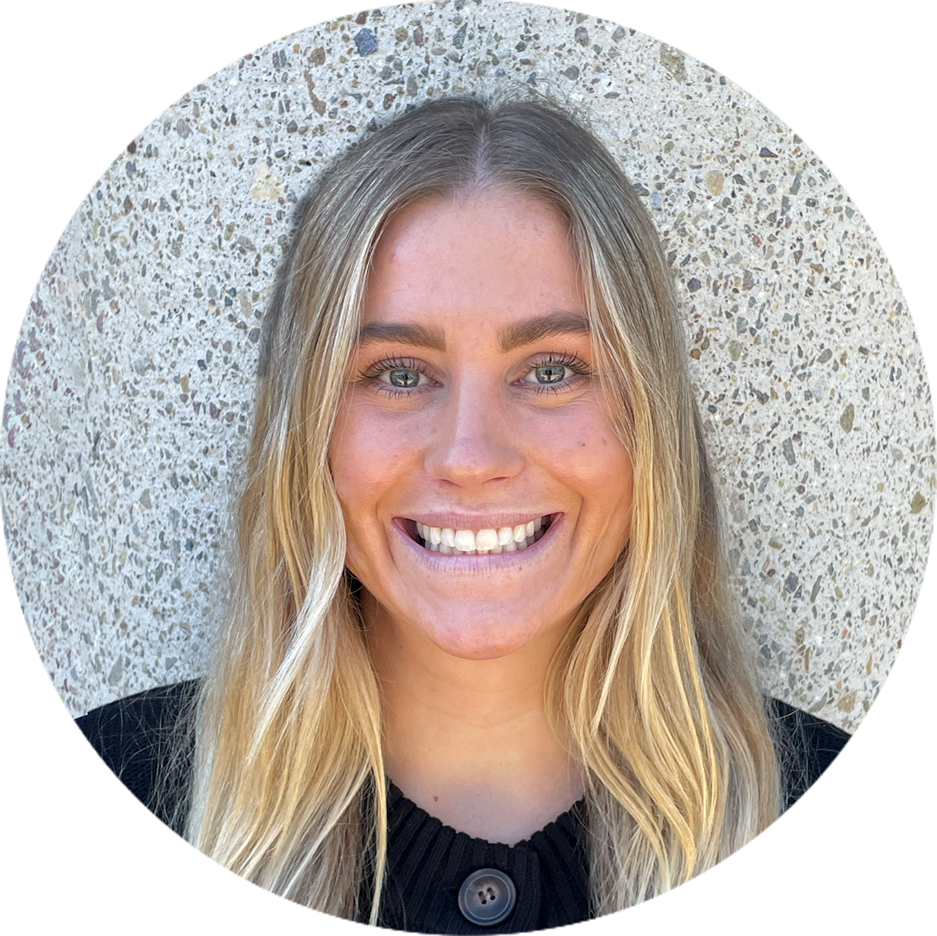 headshots-sam Smiling person with long blonde hair and a black shirt stands in front of a textured concrete wall.
