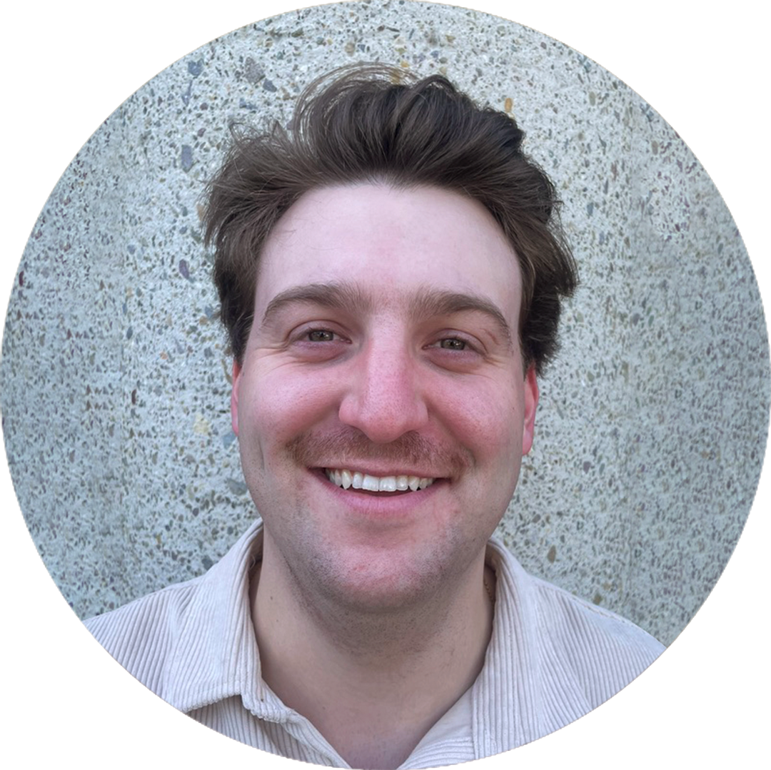 headshot-chirs-2 Person with a mustache smiling at the camera, standing against a textured stone wall.