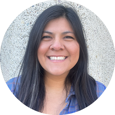 jazmin-headshots A person with long dark hair smiles while wearing a blue plaid shirt, standing in front of a textured concrete wall.
