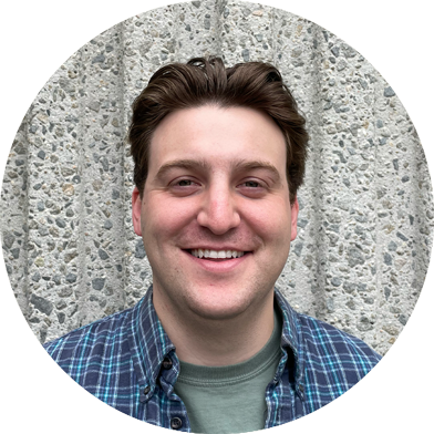chris-headshots Smiling man with short brown hair wearing a green shirt and blue plaid overshirt, standing in front of a textured concrete wall.