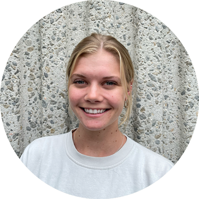 clancy-headshots A young woman with blonde hair, wearing a white shirt, smiles at the camera while standing in front of a textured concrete wall.